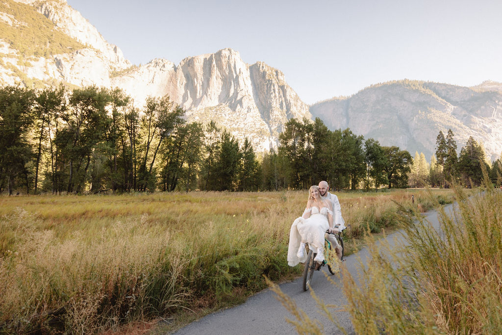 A couple rides a bicycle on a path through a grassy field with tall mountains in the background for their yosemite wedding