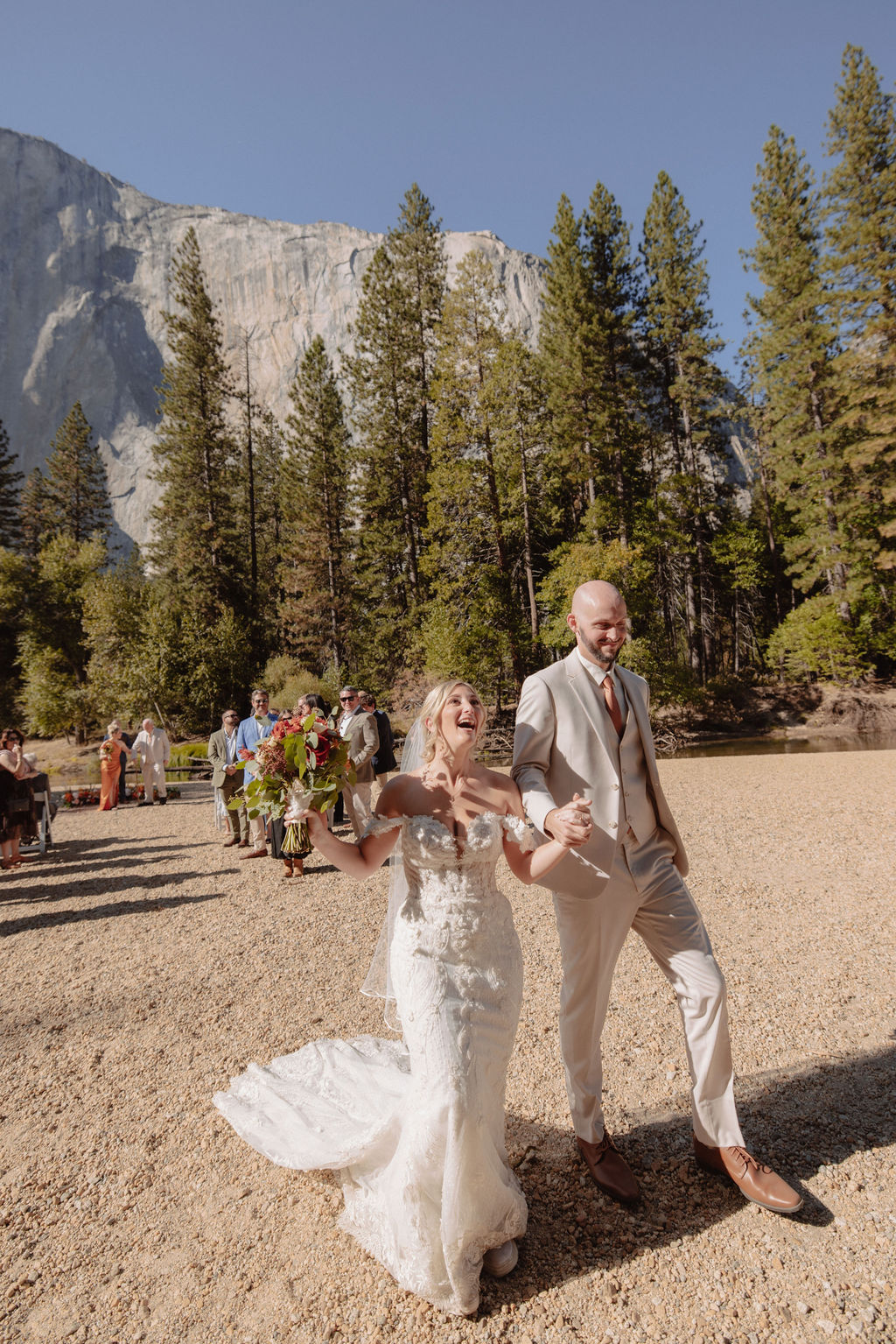 A newly married couple walks joyfully on a dirt path outdoors, with tall pine trees and a mountain in the background. Guests are visible behind them, carrying flowers.