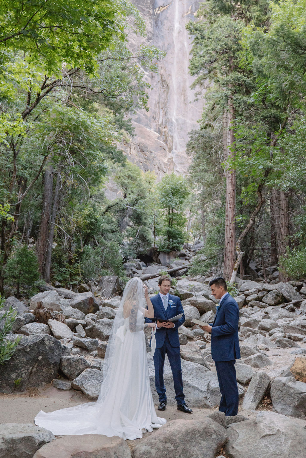 A couple stands in wedding attire on rocky terrain, with an officiant between them. Tall trees and a waterfall cascade in the background for an elopement at bridalveil falls