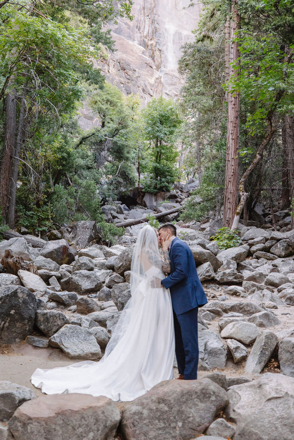 A couple stands in wedding attire on rocky terrain, with an officiant between them. Tall trees and a waterfall cascade in the background for an elopement at bridalveil falls