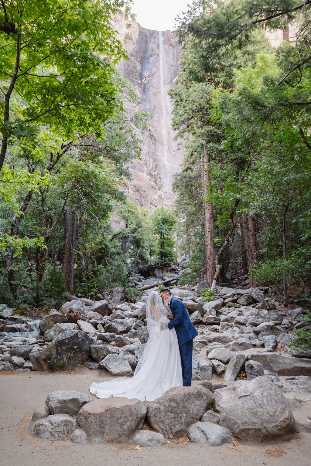 A couple stands in wedding attire on rocky terrain, with an officiant between them. Tall trees and a waterfall cascade in the background for an elopement at bridalveil falls