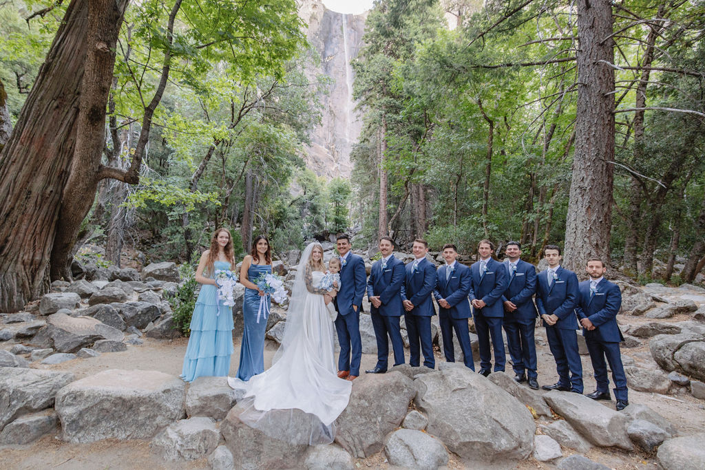 A couple stands in wedding attire on rocky terrain, with an officiant between them. Tall trees and a waterfall cascade in the background for an elopement at bridalveil falls