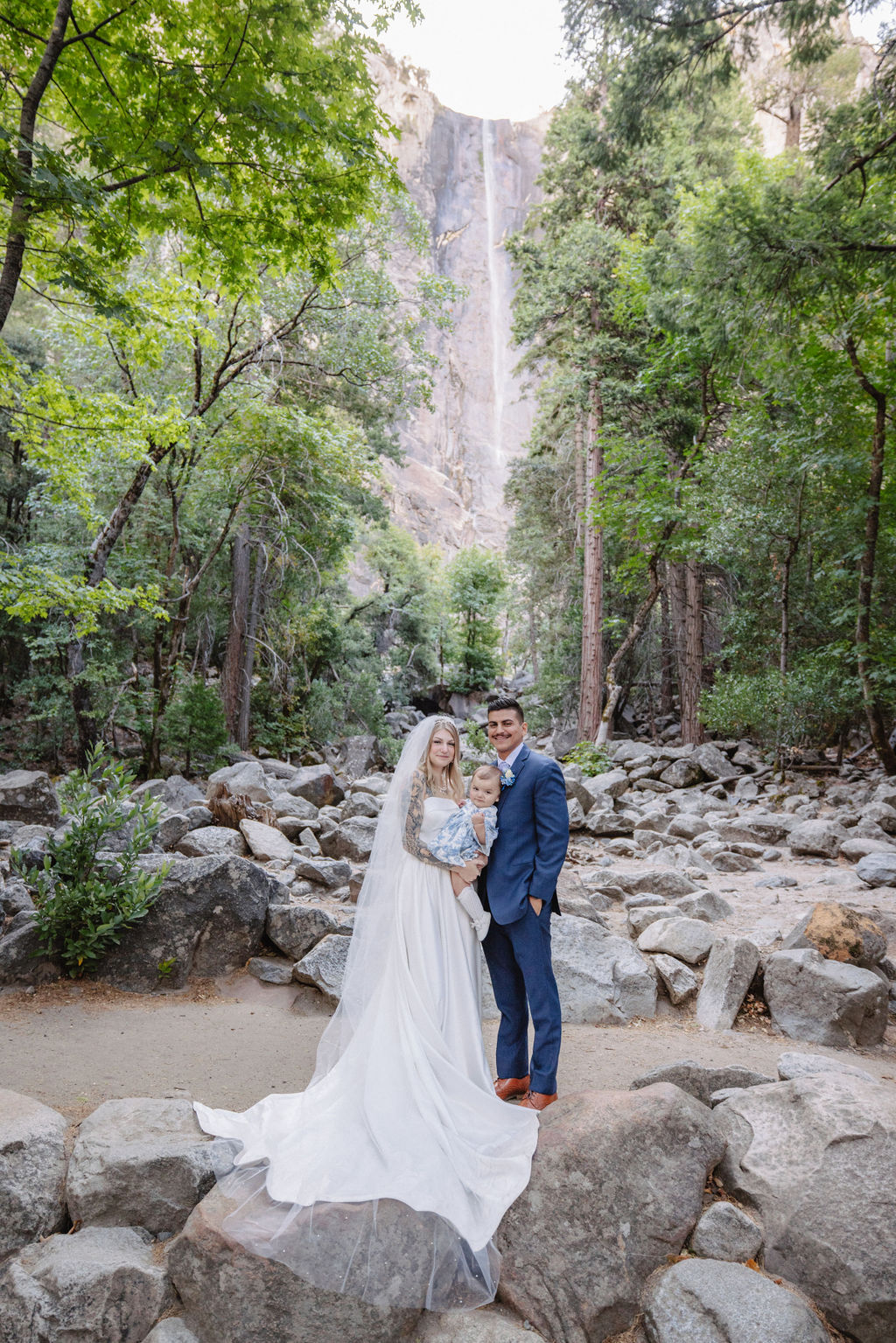 A couple stands in wedding attire on rocky terrain, with an officiant between them. Tall trees and a waterfall cascade in the background for an elopement at bridalveil falls