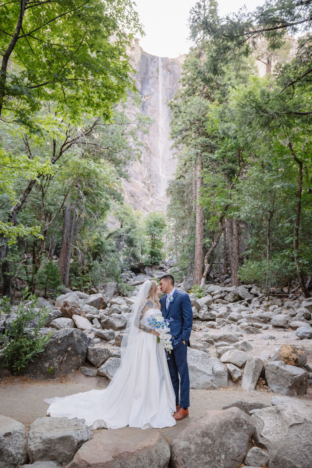 A couple stands in wedding attire on rocky terrain, with an officiant between them. Tall trees and a waterfall cascade in the background for an elopement at bridalveil falls