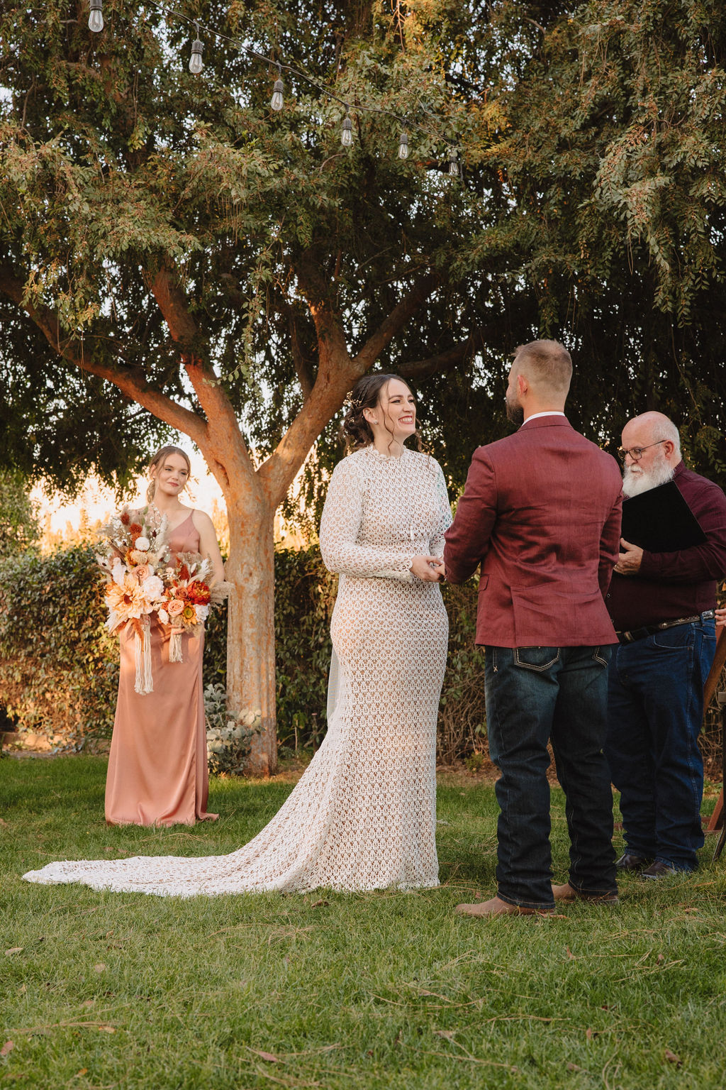 A couple is exchanging vows outdoors under a tree. The bride is in a white dress, and the groom is in a maroon jacket. A person holding a bouquet stands nearby. The Difference Between an Elopement and a Traditional Wedding