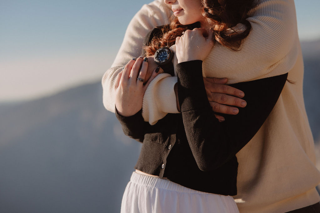 A person in a light sweater hugs someone in a dark top from behind with a scenic background ay yosemite for their candid photo shoot