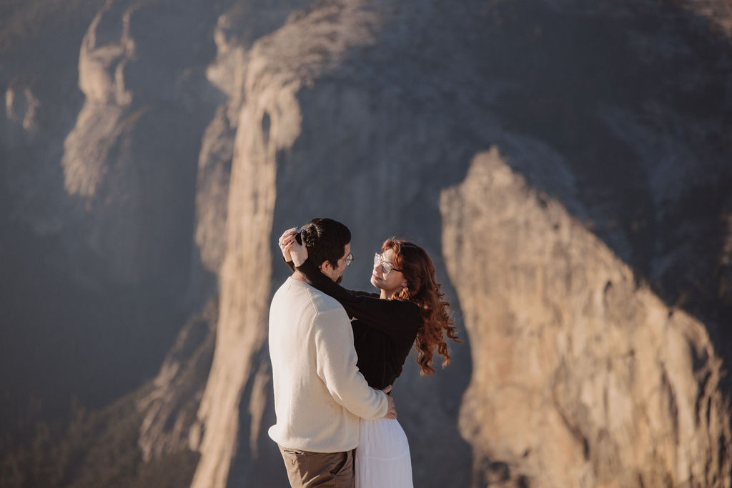 A person in a light sweater hugs someone in a dark top from behind with a scenic background ay yosemite for their candid photo shoot