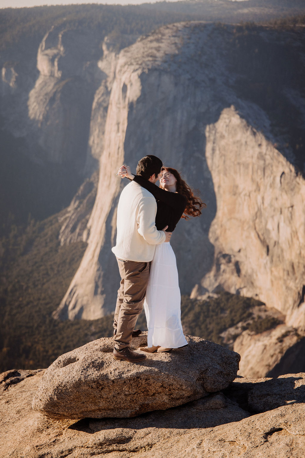 A person in a light sweater hugs someone in a dark top from behind with a scenic background ay yosemite for their candid photo shoot