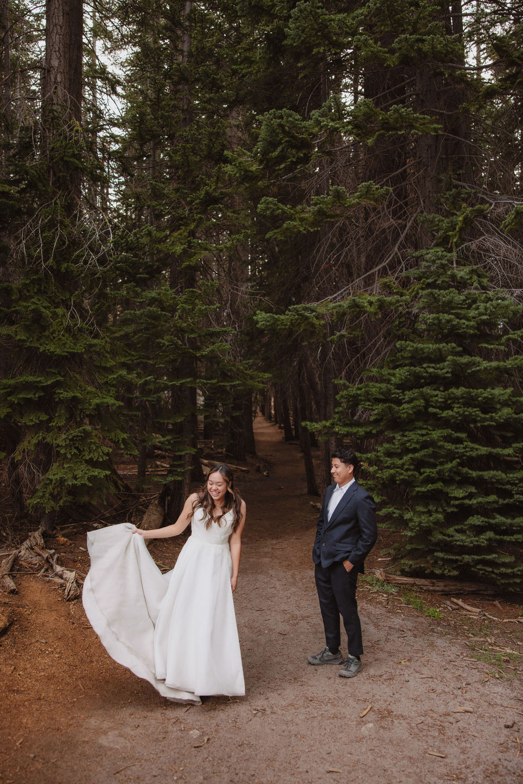 A woman in a white dress stands on a forest path, holding her dress. A man in a suit stands nearby, looking at her. Tall trees surround them for their candid photo shoot