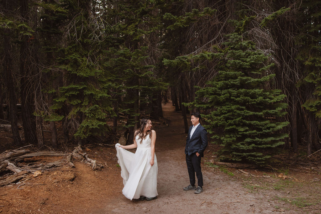 A couple stands on a forest path. The woman in a white dress looks at the man, who is wearing a dark suit. Pine trees surround them.