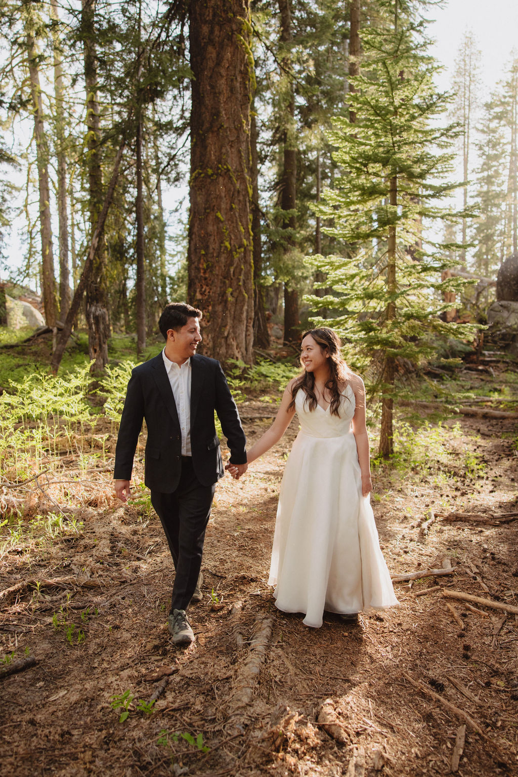 A woman in a white dress stands on a forest path, holding her dress. A man in a suit stands nearby, looking at her. Tall trees surround them for their candid photo shoot