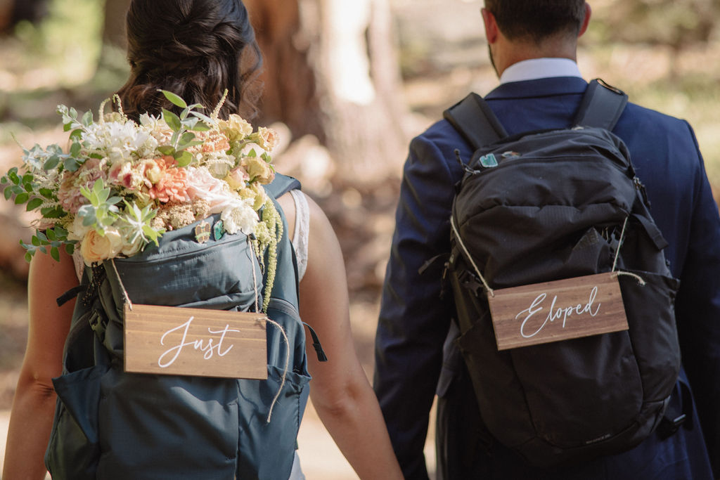 A couple is walking away, each wearing a backpack with signs reading "Just" and "Eloped" in a wooded area. The Difference Between an Elopement and a Traditional Wedding