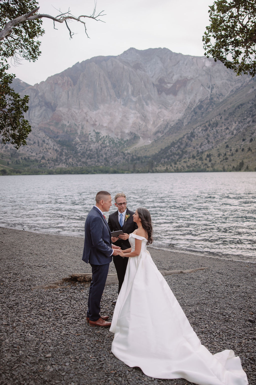 A couple in wedding attire stand with an officiant by a lake, with mountains in the background.