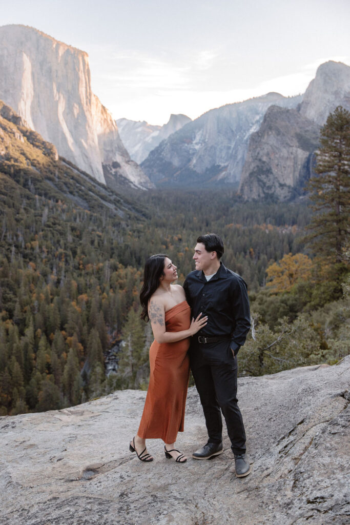 A couple stands on a rocky overlook with a scenic view of Yosemite Valley, featuring cliffs and trees in the background.