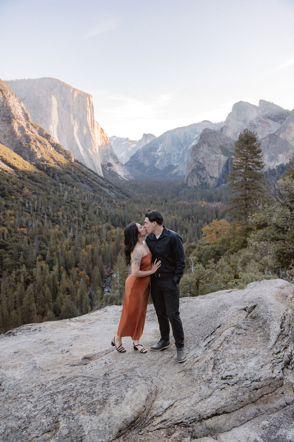 A couple stands on a rocky overlook with a scenic view of Yosemite Valley, featuring cliffs and trees in the background.