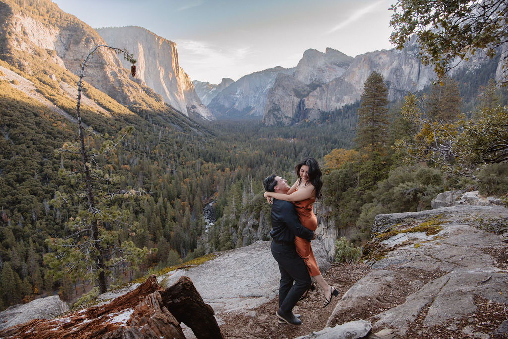 A couple stands on a rocky overlook with a scenic view of Yosemite Valley, featuring cliffs and trees in the background.