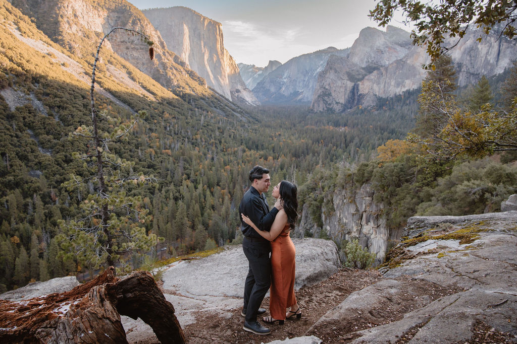 A couple stands on a rocky overlook with a scenic view of Yosemite Valley, featuring cliffs and trees in the background.