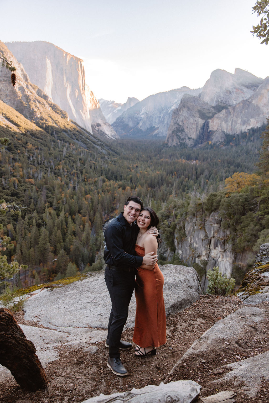 A couple stands on a rocky overlook with a scenic view of Yosemite Valley, featuring cliffs and trees in the background.