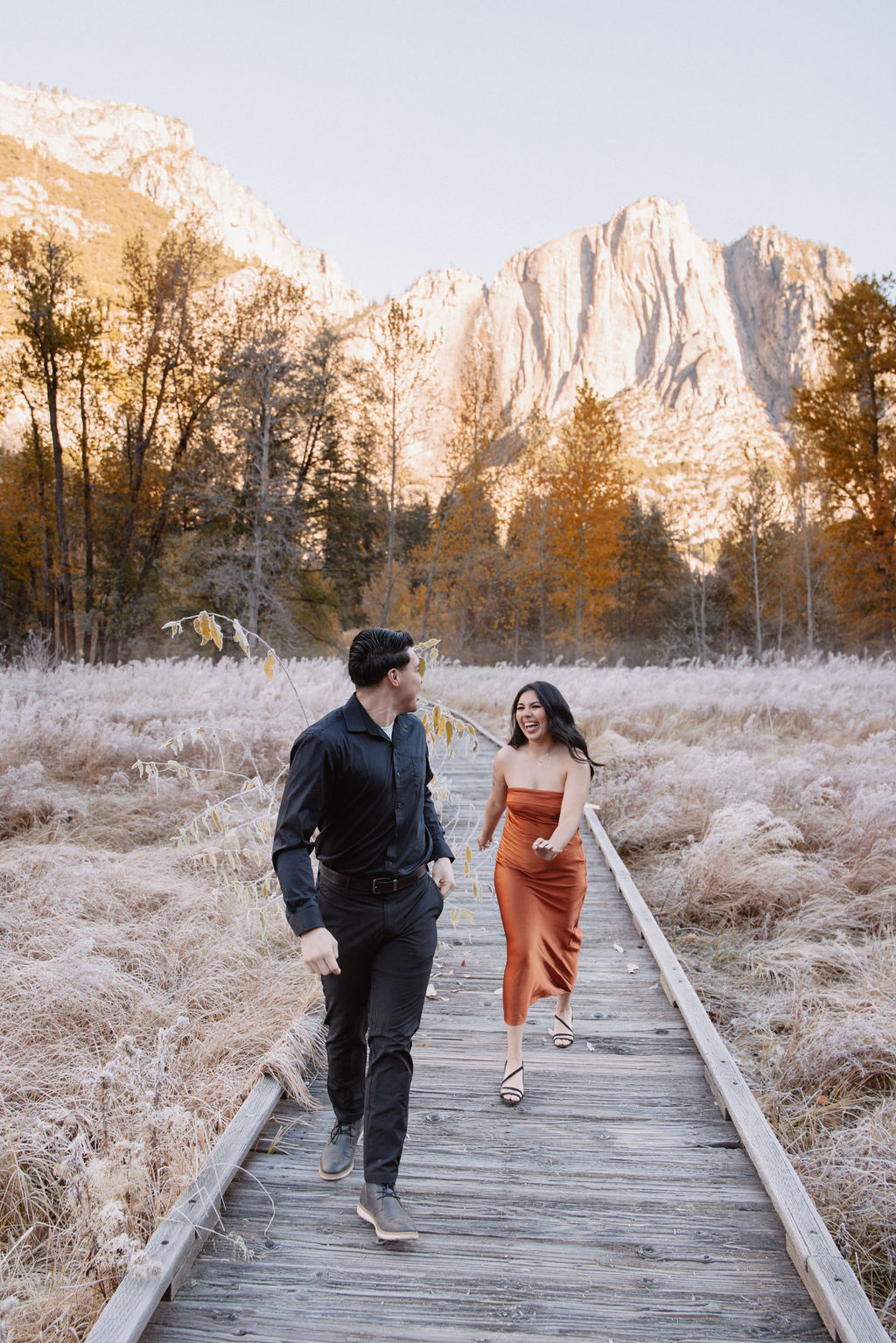 A man and woman walk on a wooden path through a grassy field with mountains and trees in the background for their yosemite fall photos