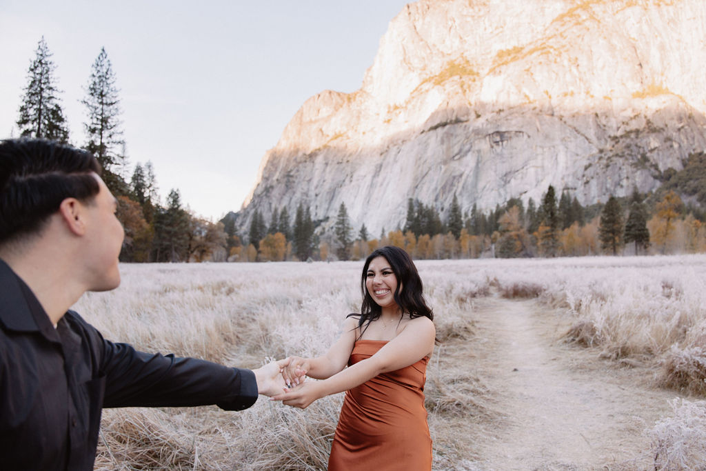 A man and woman walk on a wooden path through a grassy field with mountains and trees in the background for their yosemite fall photos