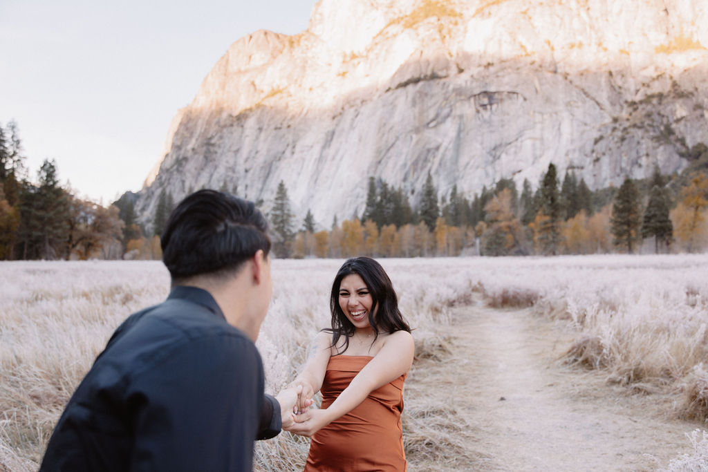 A man and woman walk on a wooden path through a grassy field with mountains and trees in the background for their yosemite fall photos