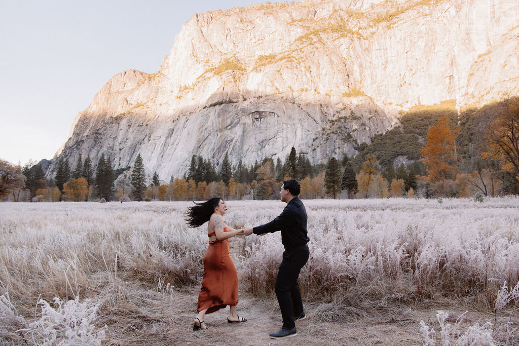 A man and woman walk on a wooden path through a grassy field with mountains and trees in the background for their yosemite fall photos