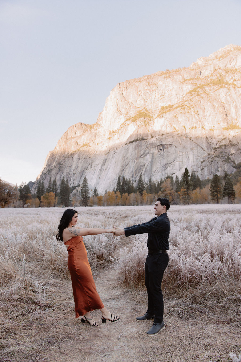 A man and woman walk on a wooden path through a grassy field with mountains and trees in the background for their yosemite fall photos