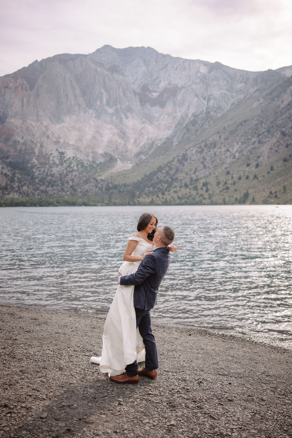 A couple in wedding attire stand with an officiant by a lake, with mountains in the background.