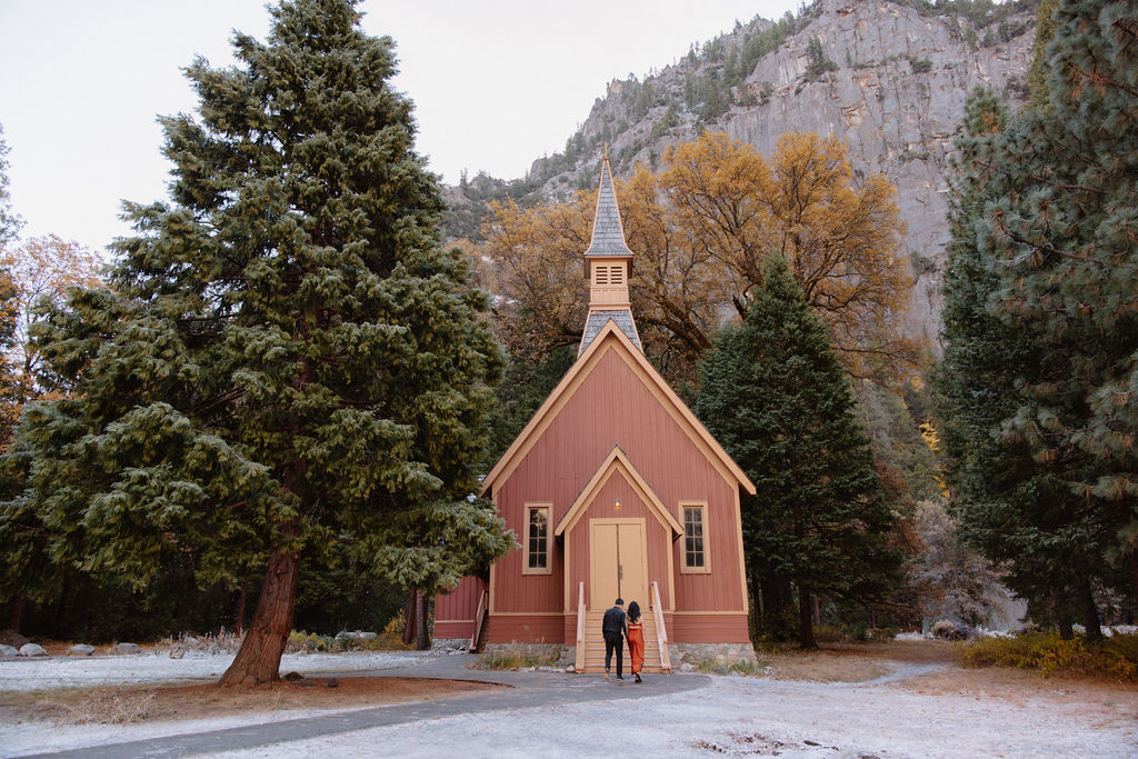 A small red church with a tall steeple, surrounded by trees and mountains. Two people stand at the entrance for their yosemite fall photos