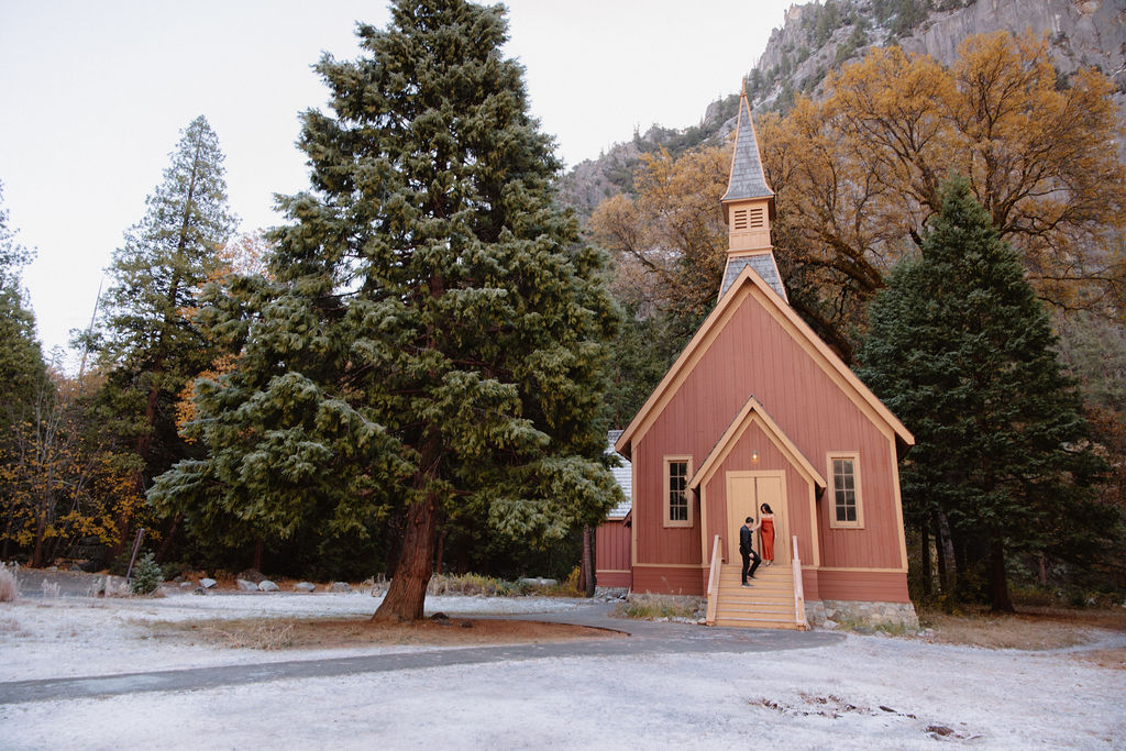 A small red church with a tall steeple, surrounded by trees and mountains. Two people stand at the entrance for their yosemite fall photos