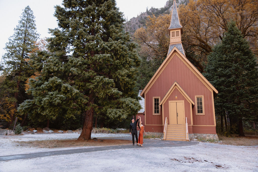 A small red church with a tall steeple, surrounded by trees and mountains. Two people stand at the entrance for their yosemite fall photos