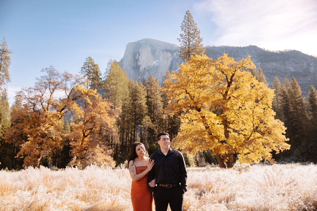 A couple stands in a scenic field with autumn trees and a mountain in the background for yosemite fall photos