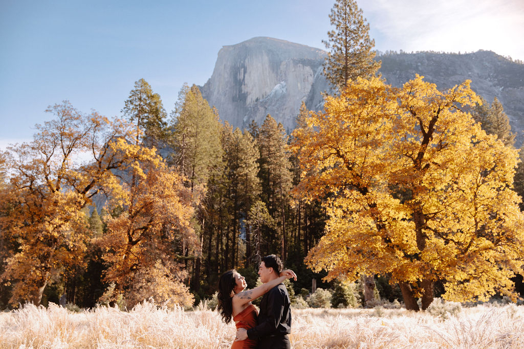 A couple stands in a scenic field with autumn trees and a mountain in the background for yosemite fall photos