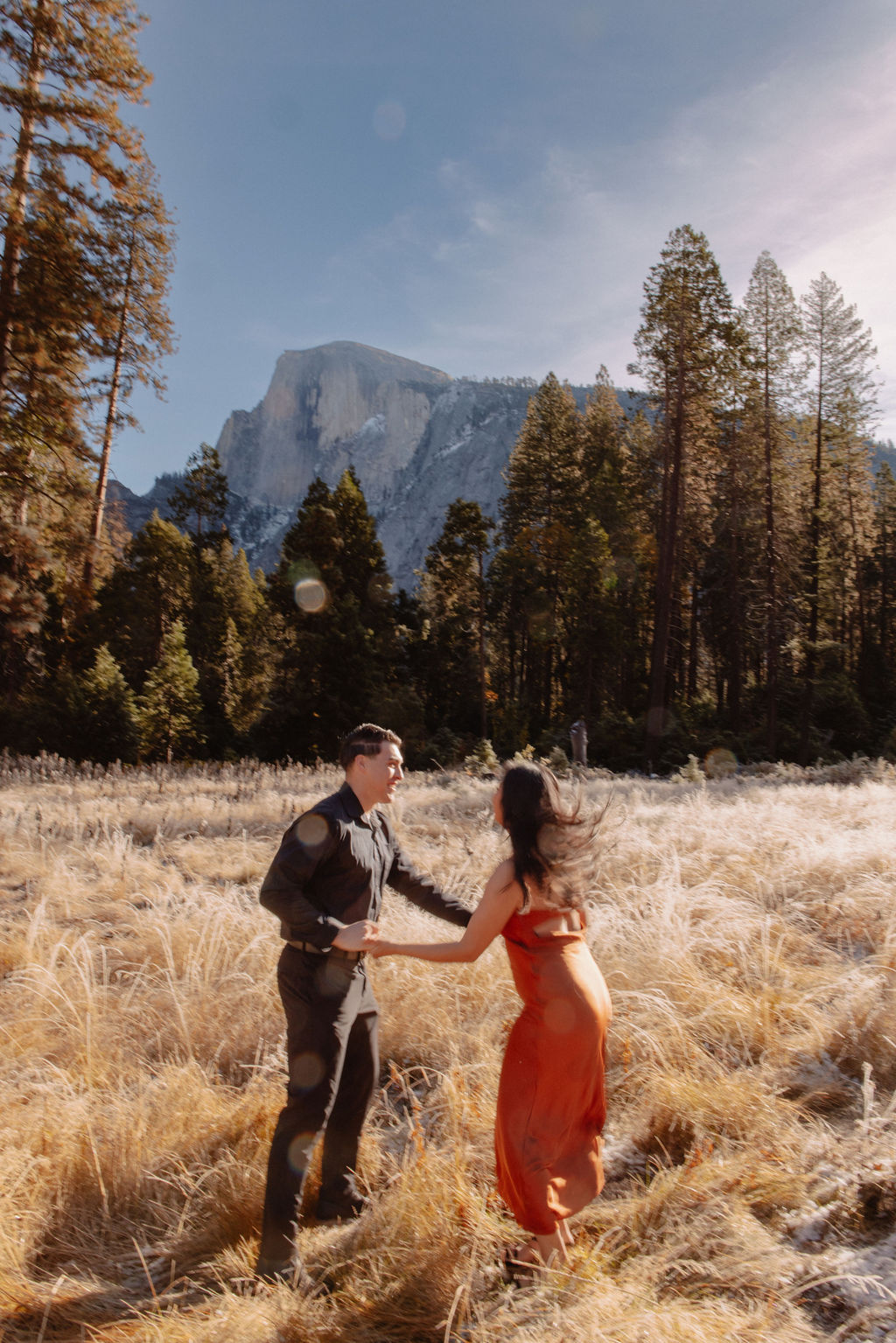 A couple walks hand in hand through a grassy meadow with tall trees and a large mountain in the background under a clear sky for their yosemite fall photos