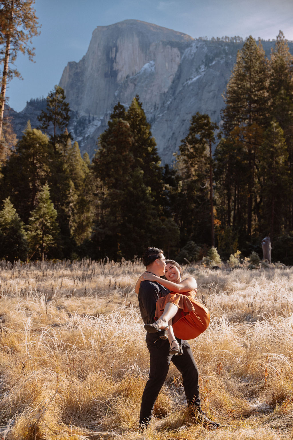 A couple stands in a scenic field with autumn trees and a mountain in the background for yosemite fall photos