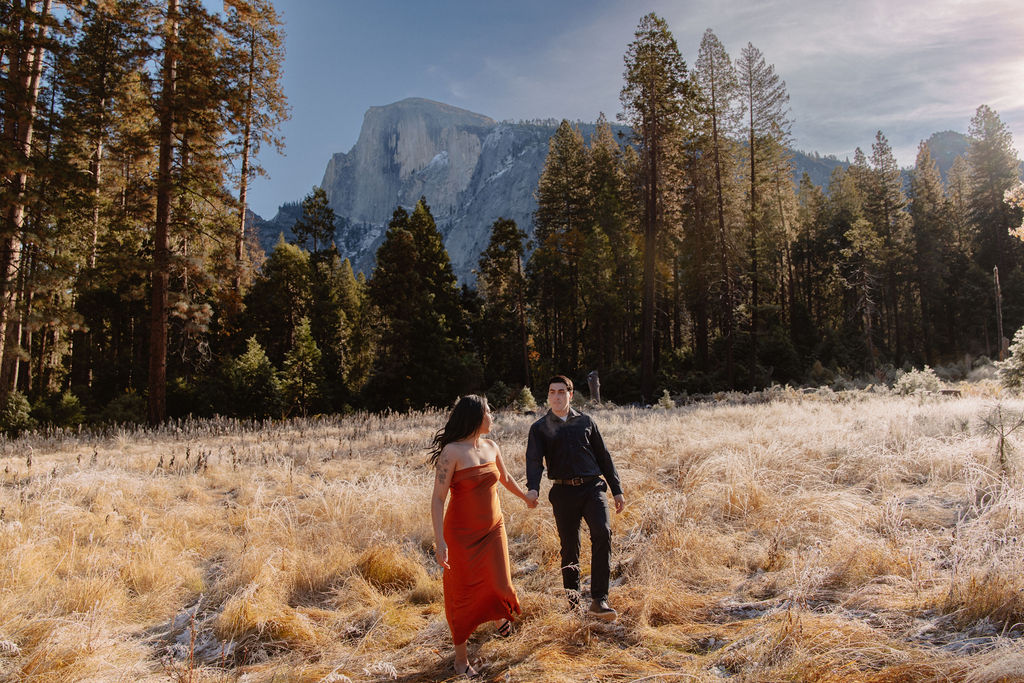 A couple walks hand in hand through a grassy meadow with tall trees and a large mountain in the background under a clear sky for their yosemite fall photos