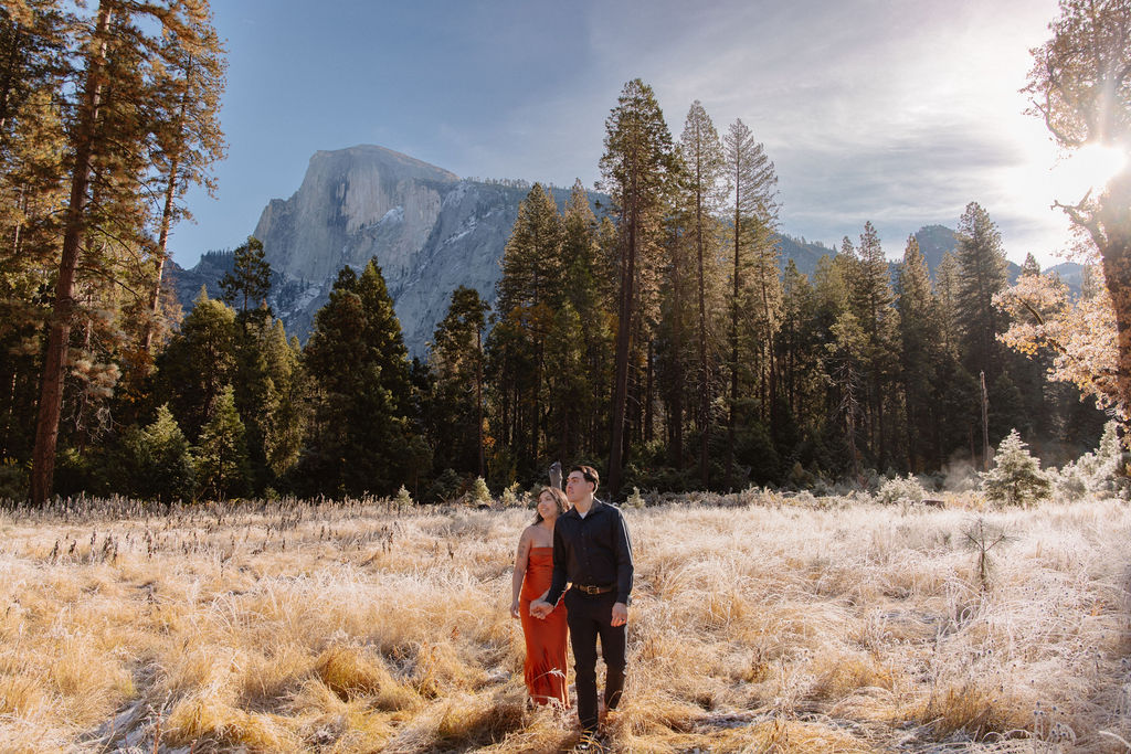 A couple walks hand in hand through a grassy meadow with tall trees and a large mountain in the background under a clear sky for their yosemite fall photos