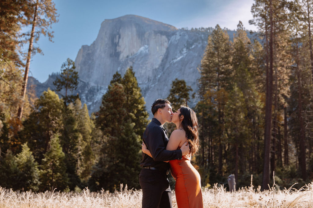 A couple walks hand in hand through a grassy meadow with tall trees and a large mountain in the background under a clear sky for their yosemite fall photos