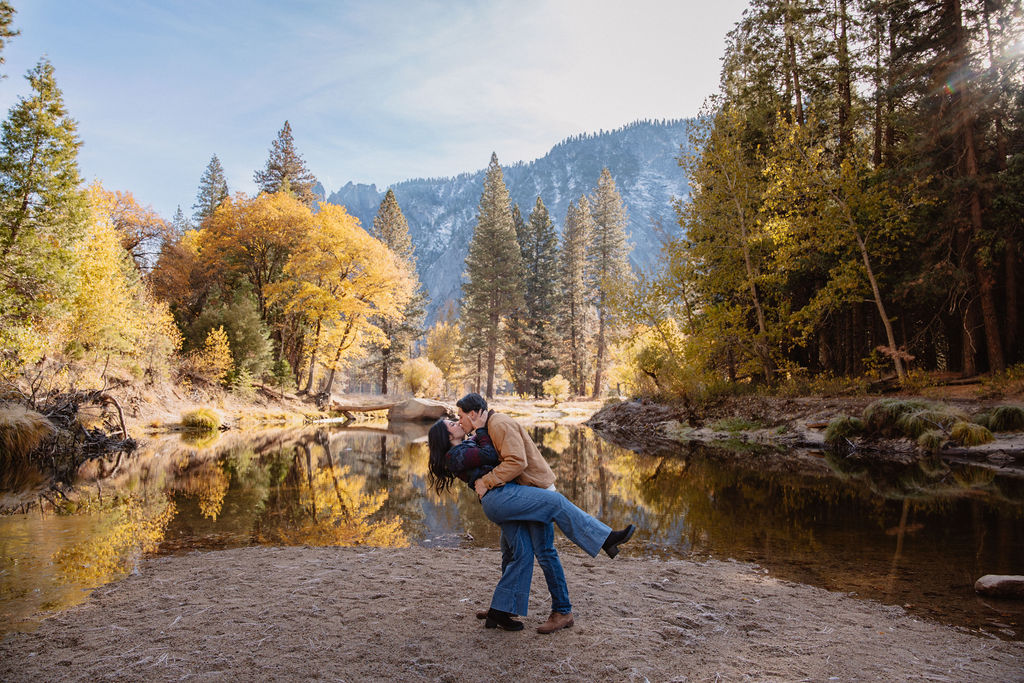 A person dips another person in an embrace by a riverbank surrounded by autumn trees and mountains for their yosemite fall photos