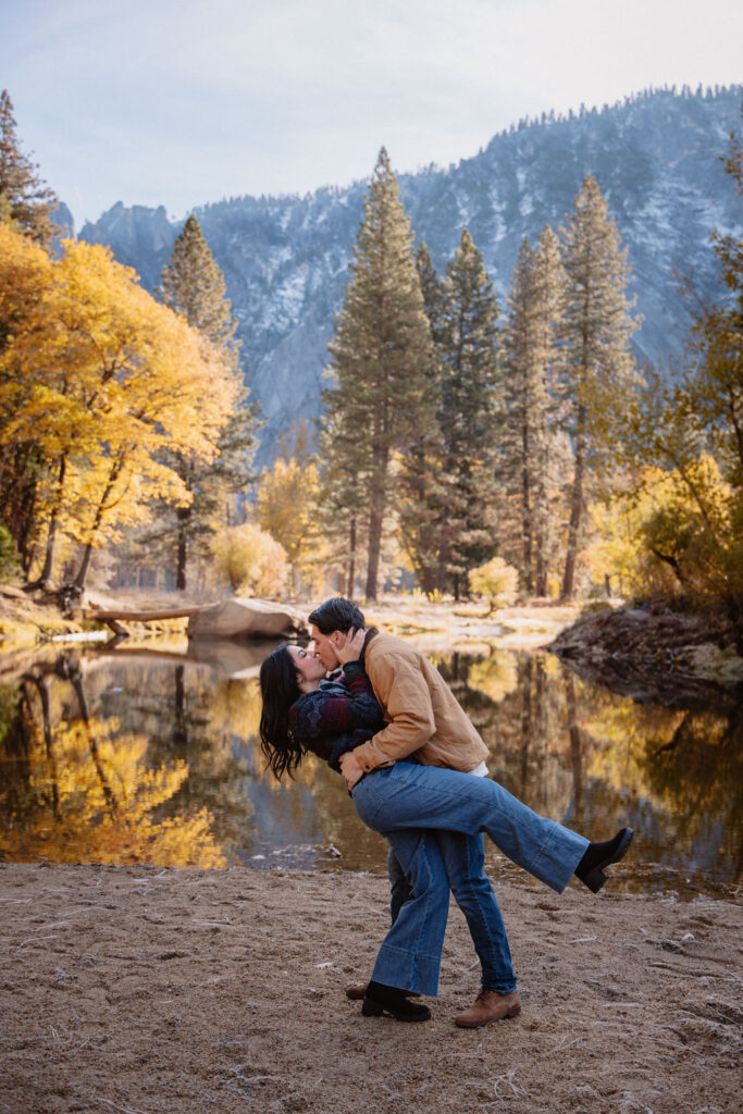 A couple kisses by a reflective lake, surrounded by trees with autumn colors and mountains in the background.