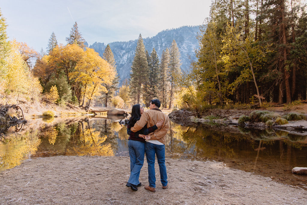 A person dips another person in an embrace by a riverbank surrounded by autumn trees and mountains for their yosemite fall photos