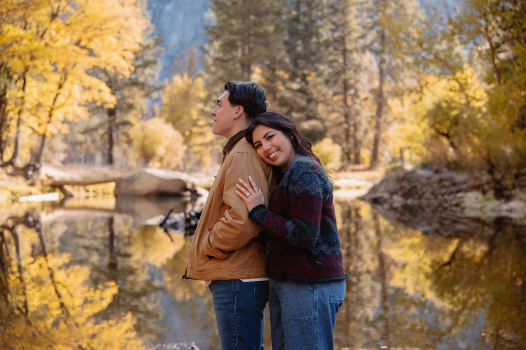 A couple kisses by a reflective lake, surrounded by trees with autumn colors and mountains in the background.