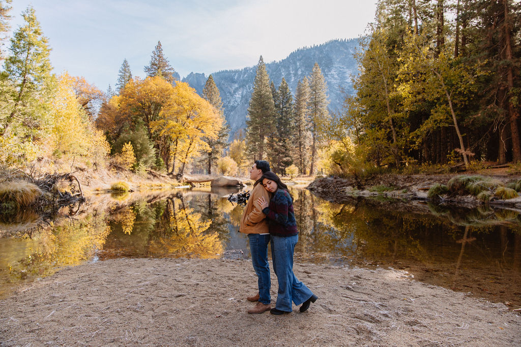 A couple kisses by a reflective lake, surrounded by trees with autumn colors and mountains in the background.