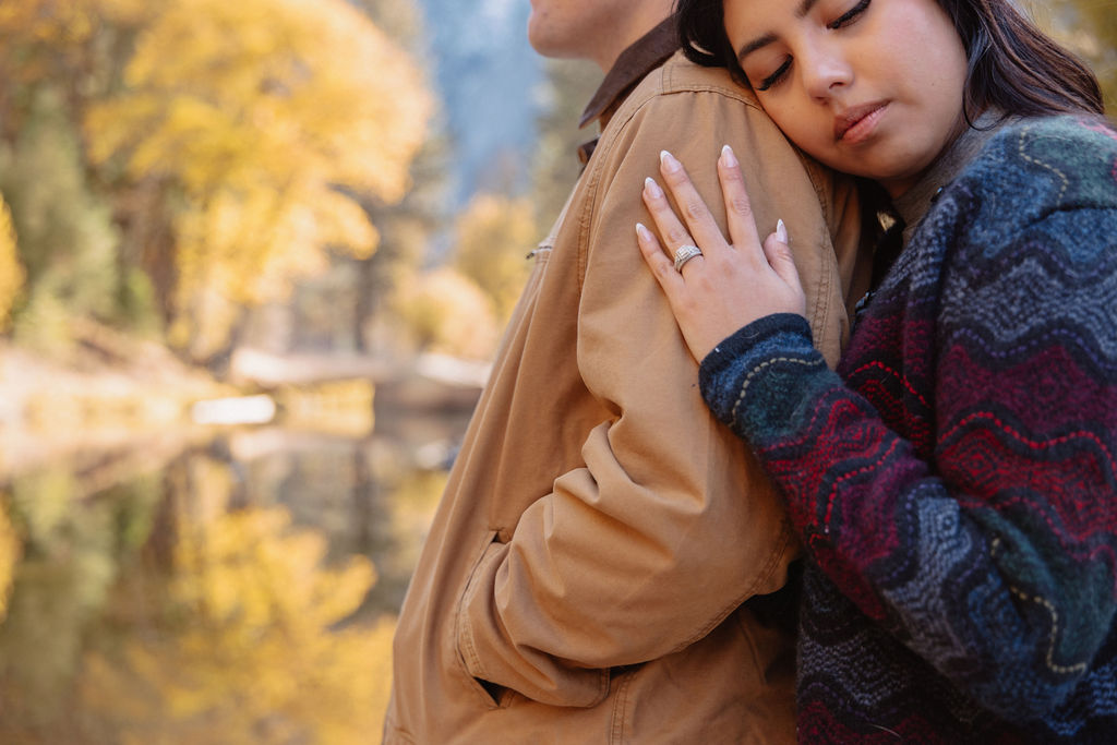 A couple kisses by a reflective lake, surrounded by trees with autumn colors and mountains in the background.