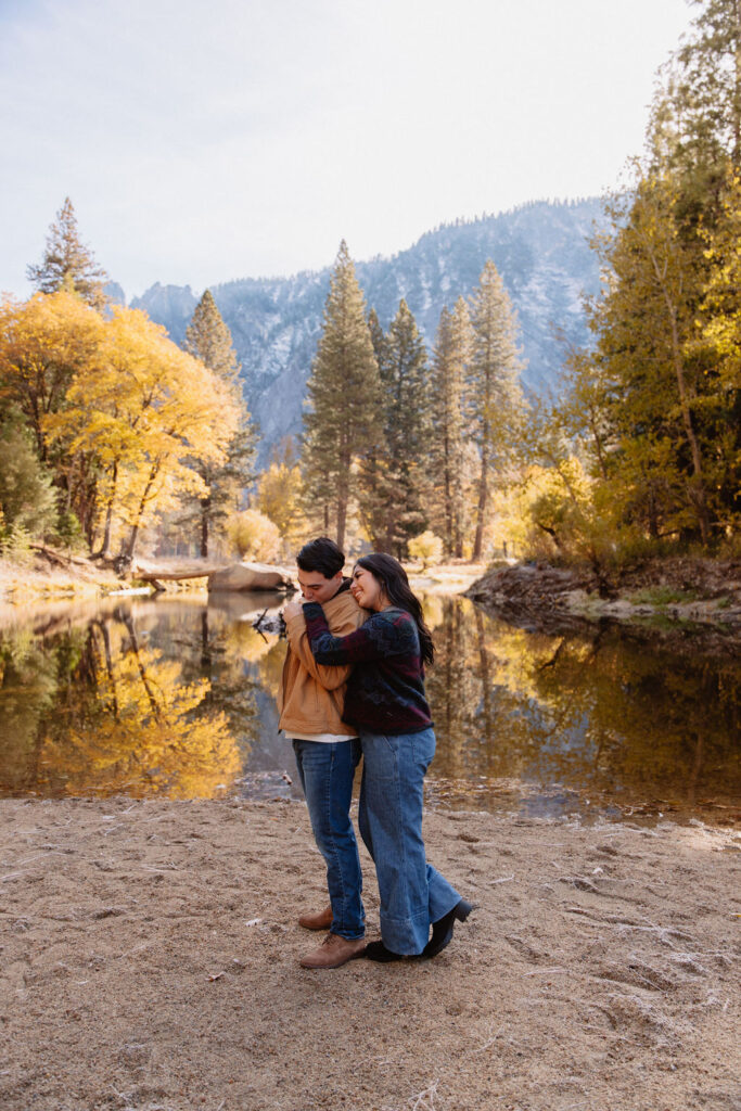 A couple kisses by a reflective lake, surrounded by trees with autumn colors and mountains in the background.