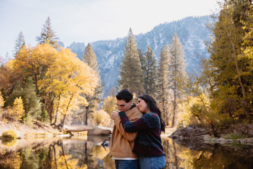 A couple kisses by a reflective lake, surrounded by trees with autumn colors and mountains in the background.