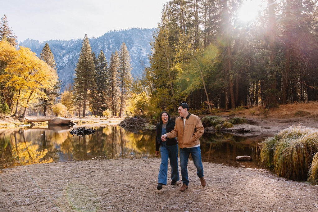 A couple kisses by a reflective lake, surrounded by trees with autumn colors and mountains in the background.