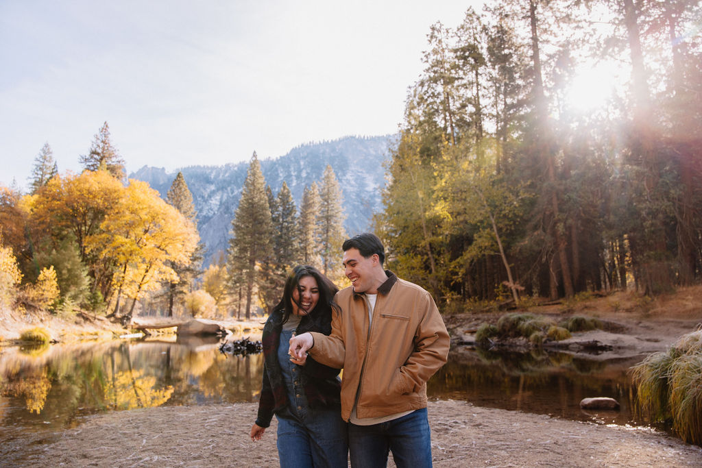 A couple kisses by a reflective lake, surrounded by trees with autumn colors and mountains in the background.