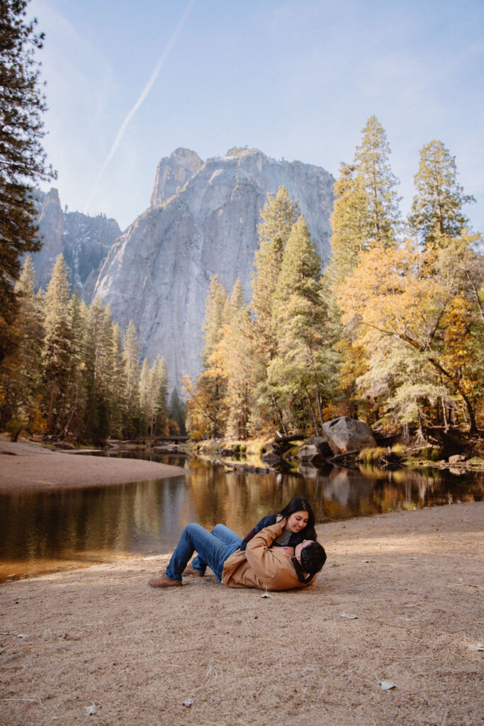 A couple kisses by a reflective lake, surrounded by trees with autumn colors and mountains in the background.