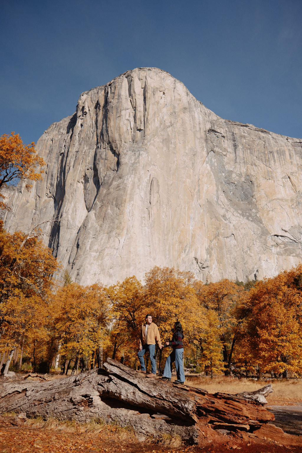 A couple kisses by a reflective lake, surrounded by trees with autumn colors and mountains in the background.